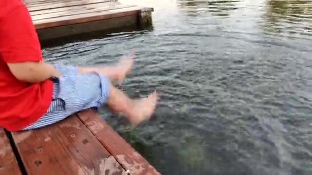 Menino está sentado na ponte e salpicando na água. — Vídeo de Stock