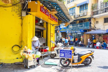 HANOI, VİETNAM 2024 Ağustos: Vietnamlı sokak satıcısı sarı deniz ürünleri dükkanının yanında konik şapkayla abur cubur satıyor
