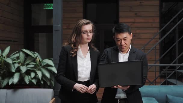 Team of two business people in formal suit Asian and a European walk through a meeting room holding a laptop with a draft business plan for a business deal with foreign partners — Αρχείο Βίντεο