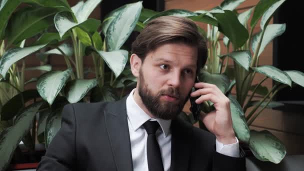 Bonito homem de negócios com barba em um terno clássico está negociando com parceiros ao telefone em um prédio de escritórios moderno em um fundo de plantas verdes. Negócios. Redes sociais. Gente. Aplicações. — Vídeo de Stock