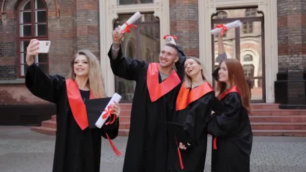 Étudiants en robes chapeaux et diplôme dans les mains prenant selfie sur fond académique. — Video