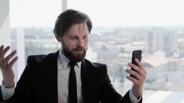 Barbudo atractivo Empresario mantener el teléfono inteligente hace una videollamada, hablar con un colega o amigo de forma remota mediante la aplicación de videoconferencia en el teléfono Big City Ventana Negocio Redes Sociales Personas Aplicaciones. — Vídeo de stock