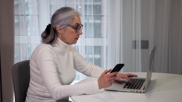 Mulher idosa moderna sentado em casa cozinha mensagens de texto no laptop usando smartphone — Vídeo de Stock