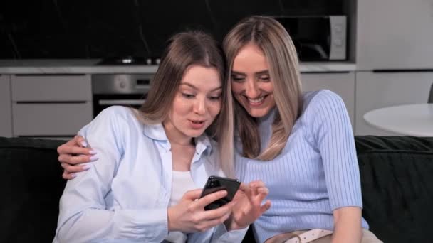 Dos lesbianas sonrientes mejores amigos abrazándose en casa y usando la aplicación en línea del teléfono. — Vídeos de Stock