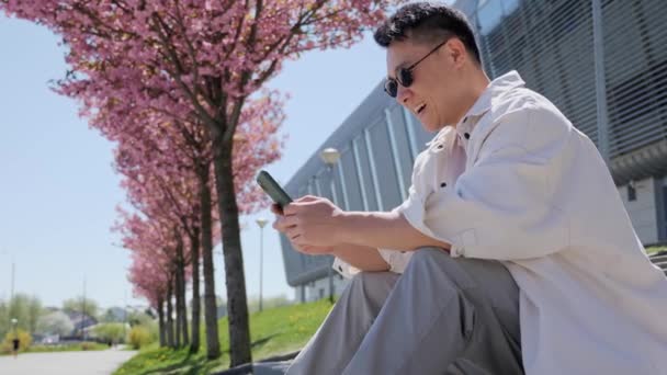 Красивий Азіатський Чат повідомлення використовуючи додаток свого смартфона на відкритому повітрі . — стокове відео