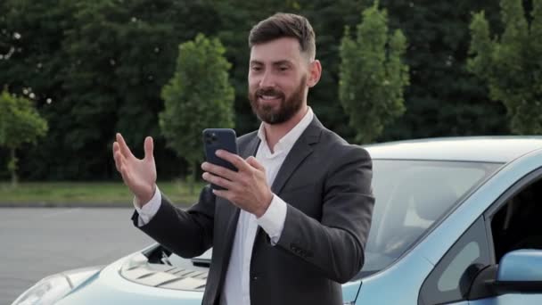 밖에 있는 새 전기 자동차 근처에서 스마트폰으로 비디오 채팅을 하는 사업가. — 비디오
