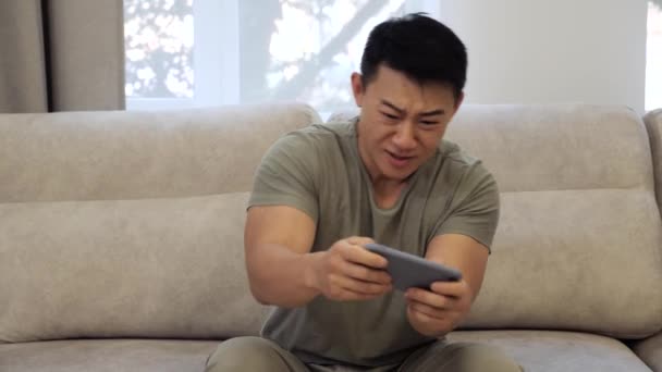 Vreugdevolle Chinese man is genieten van het spelen van video game in smartphone zitten op de bank. — Stockvideo