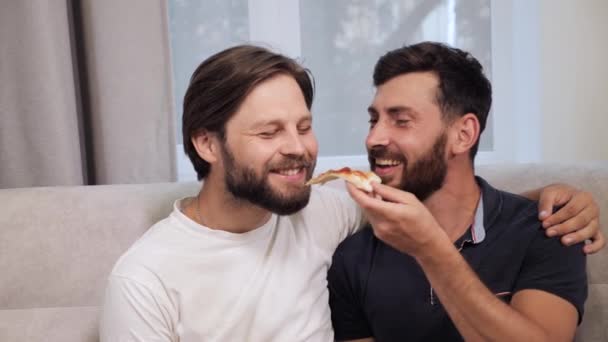 Bela feliz homens gay lgbt casal sentado no o sofá comer pizza flertando. — Vídeo de Stock