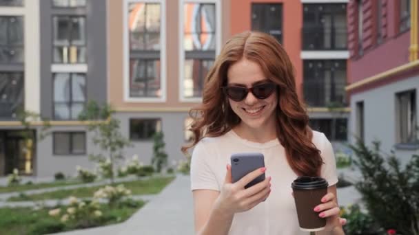 Ładna imbir dziewczyna z piegi jest chodzenie w centrum miasta trzymając telefon komórkowy. — Wideo stockowe