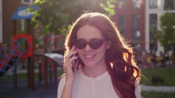 Vidám vörös hajú nő sétál végig az utcán mosolyogva beszél mobilon.. — Stock videók