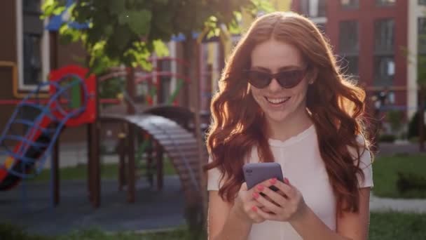 Atractiva joven pelirroja mujer caminando usando el teléfono chateando con amigos. — Vídeos de Stock