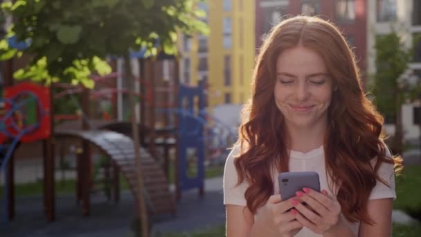 Linda mulher de cabelo vermelho digitando por telefone celular ao ar livre. — Vídeo de Stock