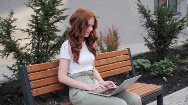 Close-up tiro de uma jovem mulher de cabelo vermelho digitando em um teclado laptop no parque. — Vídeo de Stock