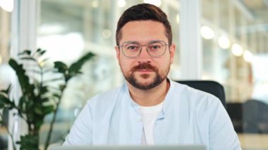 Açık mavi gömlekli, sakallı ve gözlüklü kendine güvenen iş adamı modern ofiste çalışıyor. Ciddi erkek profesyonel kameraya dostça bir ifade, başarı ve profesyonellikle bakıyor..