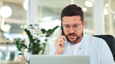 Açık mavi gömlekli ve gözlüklü neşeli iş adamı akıllı telefondan konuşuyor, modern parlak bir ofiste dizüstü bilgisayar üzerinde çalışıyor. Mutluluğu ve bağlılığı ifade eder.