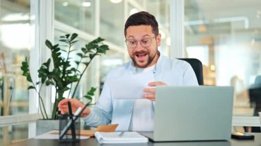 Açık mavi gömlekli ve gözlüklü hevesli bir işadamı, modern bir ofis masasında, bir mektuba heyecanla tepki veriyor, iyi haberleri kutluyor..