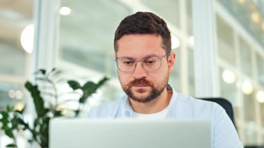 Hafif gömlek ve gözlük takan yorgun erkek işçi modern ofiste dizüstü bilgisayarla çalışıyor. Stresli uzman, ekran gerilimi ve şakak ovma yüzünden baş ağrısı çekiyor. Çok çalışan işadamı yorgun hisseder.