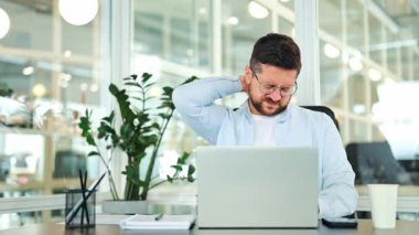 Yorgun iş adamı, açık mavi tişört, masajlar acı verici boyun. Çalışma masasında uzun saatler rahatsızlık veriyor. Modern çalışma alanı, parlak çevre. Stres ve gerginlik yüzünden anlaşılıyor. Sağlık endişesi.