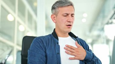 Endişeli bir adam, mavi kot gömlek ve beyaz tişört giymiş modern bir ofiste göğüs ağrısı çekiyor. Sıkıntılı ifadesi rahatsızlığı ve kalp sağlığı endişesini gösteriyor..