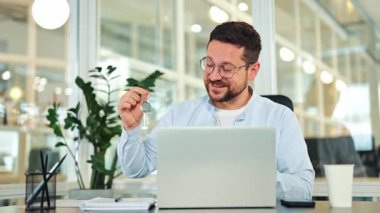 Başarılı bir iş adamı, modern bir ofiste açık mavi bir gömlek giyiyor, yeni ev anahtarlarını neşeyle sergiliyor, mülkiyet hakimiyeti hakkındaki mutluluk ve heyecanı ifade ediyor.