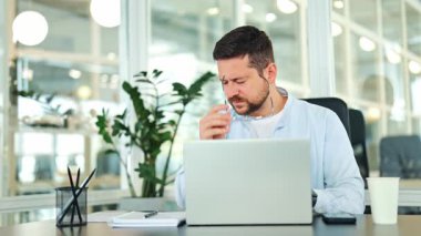 Hafif tişört giyen yorgun işadamı modern bir ofiste masa başında oturuyor. Stresli erkek, dizüstü bilgisayarla çalışırken, baş ağrısından muzdarip, bunalmış ve bitkin hissederek gözlüğünü çıkarır..