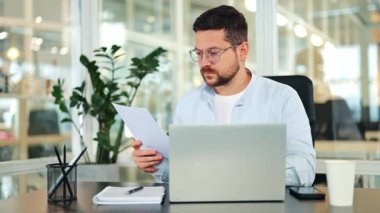 Açık mavi gömlekli ve gözlüklü odaklanmış işadamı modern bir ofis masasında dizüstü bilgisayarla belgeleri inceliyor. Profesyonel bir ortamda konsantrasyon ve ciddi çalışmalar gösteriliyor.