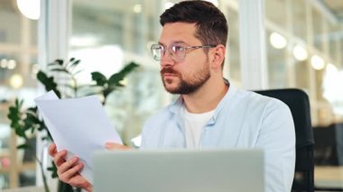 Açık mavi gömlekli ve gözlüklü düşünceli bir erkek ofis masasında belgeleri analiz ediyor. Odaklanmış işadamı, modern bir çalışma alanında derin düşünerek, bilgiyi dikkate alır..