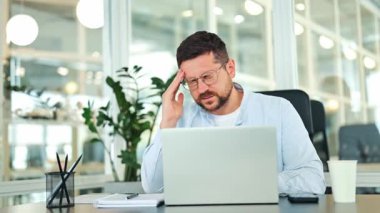 Açık mavi gömlek giyen stresli işadamı, modern bir ofis masasında dizüstü bilgisayarla çalışırken acı verici bir baş ağrısı çekiyor. Fazla çalışan uzman yorgun ve hayal kırıklığına uğramış hissediyor..