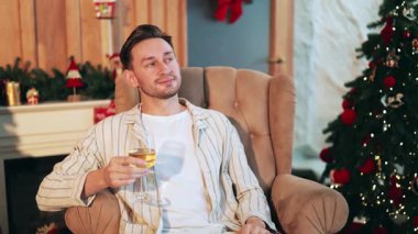 Neşeli adam elinde bardak, Noel süslemeleriyle rahatlıyor. Sıcak ışıklarla sıcacık bir tatil, huzur ve memnuniyet hissi yaratır. Kutlama sezonunu sessiz bir sevinçle kutlayın.