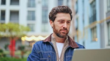 İçerik sahibi, sakallı, dışarıda dizüstü bilgisayarda özenle çalışıyor. Neşeli, kendinden emin bir gülümsemeyle, mutluluğu ve başarıyı iletiyor. Uzak çalışma, kentsel yaşam tarzı ve teknoloji