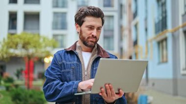 Odaklanmış, memnun bir adam canlı şehir ortamında özenle dizüstü bilgisayarla çalışır. Üretkenlik ve bağlılık ile tatmin olan, gevşek güvenle dijital görevden zevk alan.