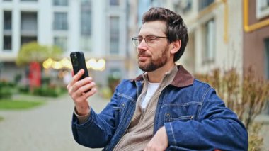 Gözlüklü adam cep telefonuyla canlı video görüşmesinden hoşlanıyor. Etkileyici bir şekilde iletişim kurar, düşüncelerini paylaşır, diğerleriyle bağlantı kurar, pozitif bir bağ hisseder. Uzak etkileşim için modern kentsel ortam