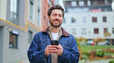 Dışarıda mutlu bir adam, akıllı telefona dalmışken gülümsüyor. Modern şehir ortamında teknolojiyle bağlantının tadını çıkarmak. İçerik ve pozitif hissetmek.