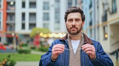 Sakallı adam güvenli bir şekilde dışarıda kameraya bakıyor. Gözlük takan, neşeli ifadeler veren, net görüşlü biri. Şehir merkezi, parlak bir gülümseme. Mutlu, zeki, memnun..