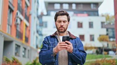 Açık havada duran, akıllı telefonla meşgul biri. Kafa karışıklığı ve endişeyi tecrübe eder, sonra tekrar cihaza bakmadan önce derin derin düşünür. Belirsizliği yansıtır.