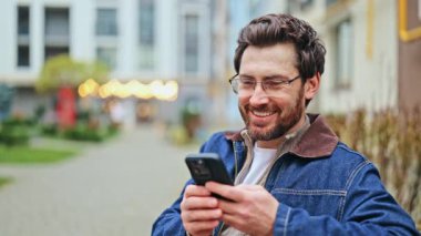 Şehir ortamında akıllı telefonla uğraşan kişi gülümser, mutlu olur. Teknoloji, neşe ve içerik anlarını birbirine bağlar. Günlük açık hava etkileşimi.