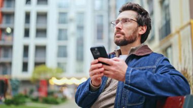 Gözlüklü sakallı adam dışarıda akıllı telefonlardan hoşlanır. Şehir ortamında modern teknolojiyle etkileşime girerken mutluluğu buluyor. Gülen yüz neşe ve bağlantı gösterir. Anın tadını çıkar..