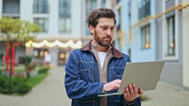 Açık bir şehir ortamında dizüstü bilgisayara yoğunlaşmak. İfade, kendini adamış odaktan memnuniyet duygusuna kayar, olumlu duyguların verimliliği ve dijital bağlantının altını çizer.