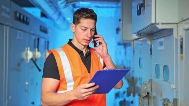 Güvenlik yeleği mühendisi panoyu incelerken telefonda konuşuyor. İşçi ciddi ve odaklanmış görünüyor, endüstriyel elektrik ortamında operasyonel görevleri yönetiyor.