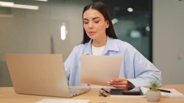 Modern ofisteki bilişim uzmanı kadın bilgisayarla uğraşırken belgelere bakıp gülümsüyor. Kariyerinde mutlu ve kendinden emin. Gösteriler başarılı iş ortamı ve üretkenlik.