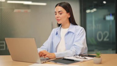 Genç iş kadını özenle dizüstü bilgisayarda çalışıyor. Görevleri tamamlarken, kendine güvenen ve olumlu bir gülümseme ortaya çıkar. Kendini işine adamış profesyonel, modern bir ofiste başarılı oluyor..