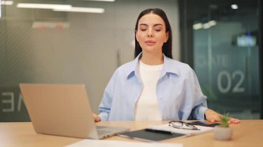 Modern ofisteki genç iş kadını gülümsüyor, kendine güveni ve ulaşılabilirliği yüksek. Dizüstü bilgisayarlı, aksesuarlı profesyonel bir ortam. Sakin, hoş geldin ve olumlu bir tutum sergiliyor..