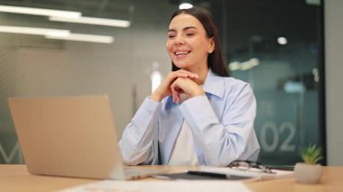 Genç iş kadını, özgüven ve olumlu bir coşku sergileyerek dizüstü bilgisayarında sanal bir toplantı yapıyor. Uzaktan iletişim kurmak ve gülümsemek, önemli konuları tartışırken doğal olarak jestler yapar..