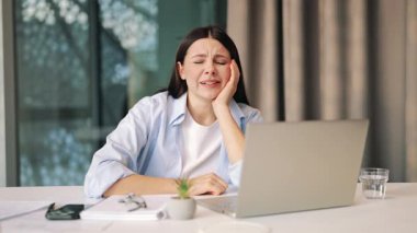 Genç bir kadın ofis masasında dizüstü bilgisayarla çalışırken yorgun ve uykulu hissediyor. İfadeler stresi, hayal kırıklığını ve aşırı çalışmanın muhtemel baş ağrısını gösteriyor. Geçerli görevle ilgili yorucu ve rahatsızlık verici.