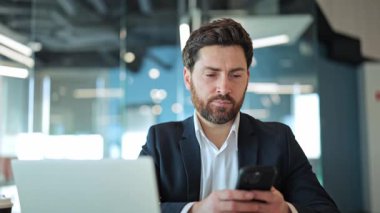 İş yerinde kendine güvenen işadamı kameraya bakıyor, teknolojiyle mutlu mesut ilgileniyor, yukarı bakıyor ve gülümsüyor. Modern ofis ortamında profesyonellik, başarı ve pozitifliği yansıtır..