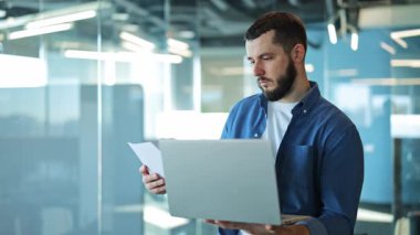 Gösterişli bir ofiste çalışkan bir profesyonel, önemli belgeleri dikkatle incelerken bir dizüstü bilgisayarla uğraşır. Odaklanmış ifadesi, konsantrasyonu ve görevlere bağlılığı iletir..