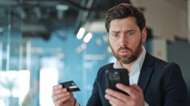 İş adamı, kredi kartı ve akıllı telefonuyla online ödeme yapmaya çalışırken şok olmuş ve endişeli görünüyor. Kafası karışmış, şaşırmış ve beklenmedik alım satım sorunları yüzünden hayal kırıklığına uğramış..