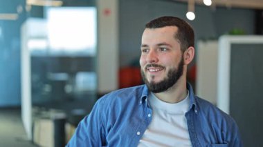 Gülümseyen profesyonel bir adam modern ofis ortamında rahatça dinlenir. Memnuniyeti, memnuniyeti, yoğun bir günde kısa süreli sakinliği yansıtır. Başarılı iş-yaşam dengesi.