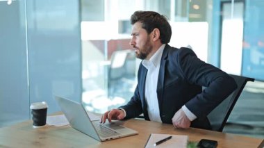 Odaklanmış iş adamı ofis masasında uzun saatler boyunca yoğun sırt ağrısı hisseder. Uzun süren dizüstü bilgisayar kullanımından kaynaklanan kronik duruş sorunları gibi rahatsızlıklar ortaya çıkıyor. Acı ve yorgunluk, rahatlama arayışı..