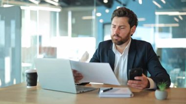 Profesyonel adam belgeleri dikkatle inceler, akıllı telefon ve dizüstü bilgisayardaki bilgileri karşılaştırır. Ticari görevlere ve modern işyeri verimliliğine ciddi bir şekilde odaklandığını gösteriyor..